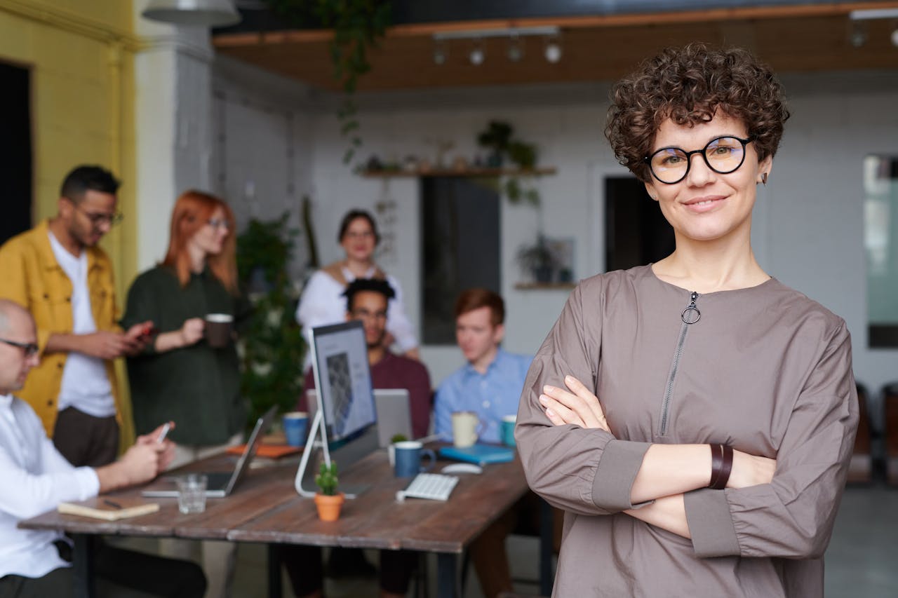Home Smiling businesswoman with curly hair stands confidently in a modern office space with colleagues.